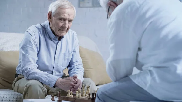 grey haired man thinking near chessboard and blurred friend at home