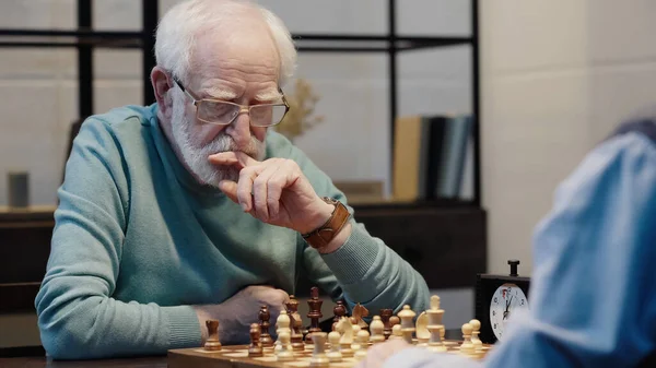 pensive man holding hand near face while playing chess with senior friend
