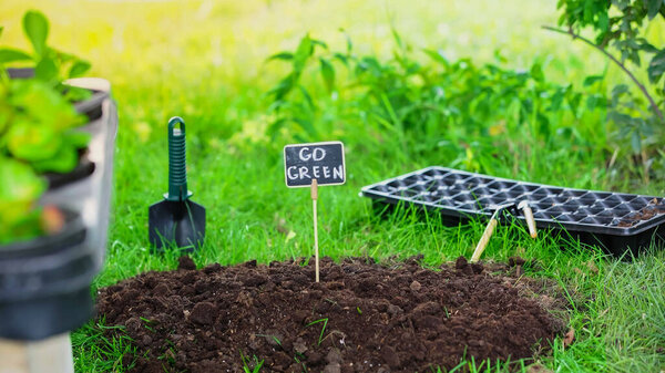 Board with go green lettering in soil near gardening tools and grass in garden 