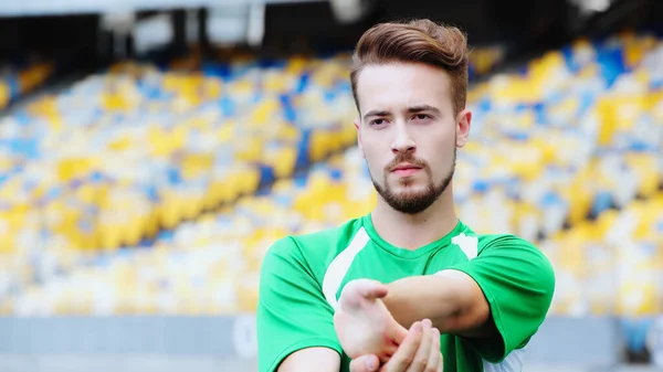 football player in green t-shirt stretching hands while warming up on stadium