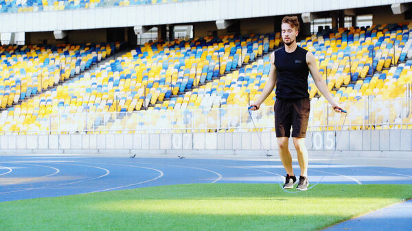 full length of bearded sportsman jumping with skipping rope on green grass at stadium 