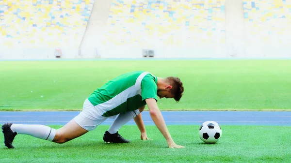 side view of football player stretching legs on green grass bear soccer ball 