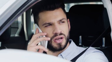 young bearded man driving car while looking away and talking on smartphone