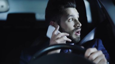 shocked businessman talking on smartphone and looking aside while driving car at night