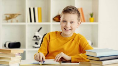 happy schoolboy doing homework near smartphone and books on desk