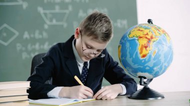 schoolboy in glasses writing in notebook near globe and books on desk