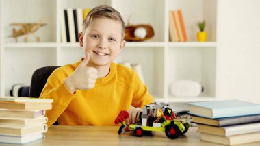 happy schoolboy sitting near toy car made of building blocks and showing thumb up at home 