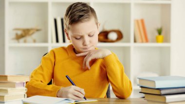 thoughtful preteen schoolboy writing in notebook near books on desk 