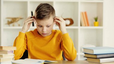 tensed preteen boy looking at notebook while doing homework near books on wooden desk 