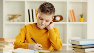 thoughtful preteen schoolkid writing in notebook while doing homework near books on wooden desk