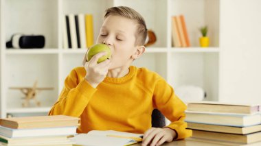 schoolboy with closed eyes biting green and tasty apple near books at home 