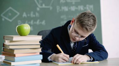 schoolboy in glasses writing in notebook near apple on books in classroom 