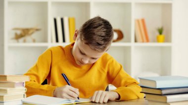 preteen boy writing in notebook while doing homework near books on wooden table at home