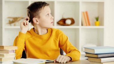 pensive preteen boy thinking while holding pen and doing homework near books on wooden table