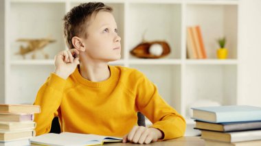 pensive preteen boy thinking while doing homework near books on wooden table at home