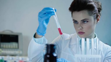 pretty scientist looking at red liquid in test tube near blurred medical equipment 