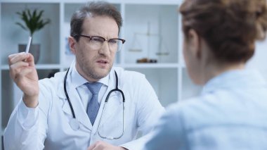 serious doctor in glasses holding cigarette while talking to blurred woman in hospital