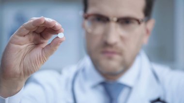 blurred doctor in glasses and white coat looking at camera and holding pill 