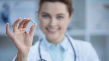 blurred and cheerful doctor in white coat smiling while holding capsule 