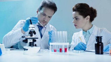 scientist taking sample with pipette near colleague making notes in lab