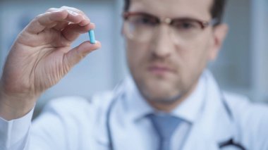 blurred doctor in glasses and white coat looking at camera and holding capsule