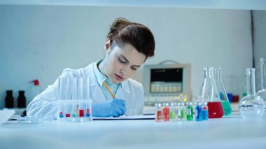 pretty scientist in white coat writing down result near test tubes and flasks with liquid in laboratory