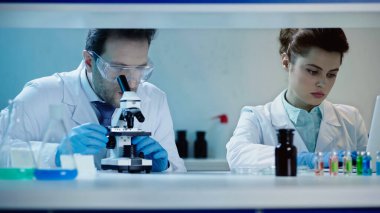 scientist in goggles looking through microscope near colleague using laptop in laboratory
