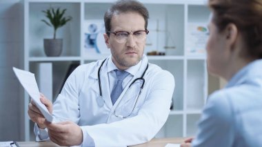 cardiologist in glasses and white coat holding cardiogram and talking to woman in clinic 