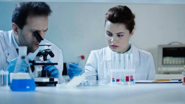 scientist in white coat looking through microscope near coworker in laboratory  