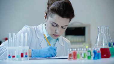 attentive female scientist writing on clipboard result of analysis near test tubes  