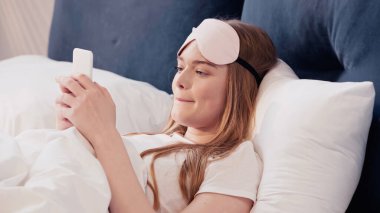 Young woman in sleep mask using smartphone on bed in morning 