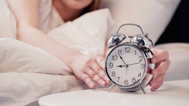 Cropped view of blurred woman touching alarm clock in bedroom in morning 