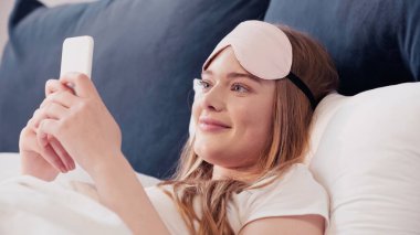 Cheerful woman in sleep mask using cellphone on bed at home 