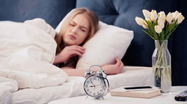 Displeased woman looking at alarm clock near smartphone in bedroom 