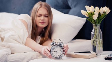 Upset woman touching alarm clock near flowers and smartphone in bedroom 