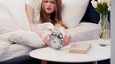 Displeased woman touching alarm clock near book and flowers in bedroom in morning  clipart