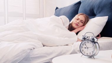 Sad woman looking at alarm clock on bedside table in bedroom clipart
