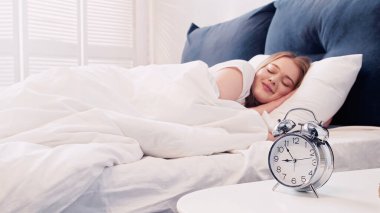 Smiling woman lying on bed near alarm clock in morning 