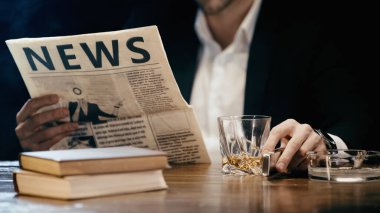 Cropped view of blurred businessman reading newspaper near whiskey and books on table isolated on black 