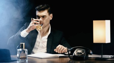 Businessman drinking whiskey near telephone and notebook on black background with smoke 