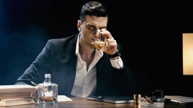 Businessman drinking whiskey near notebook and books on table on black with smoke 