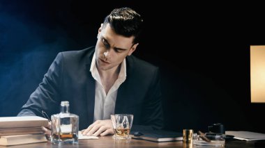 Businessman writing on notebook near whiskey and ashtray on black background with smoke 