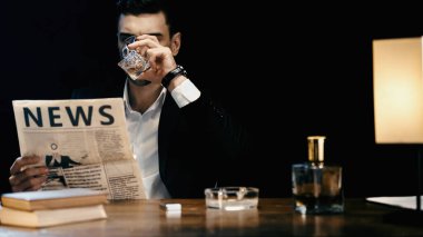 Businessman drinking whiskey and reading newspaper near books and ashtray on table isolated on black 