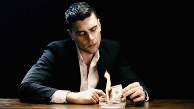 Businessman holding burning money near ashtray isolated on black 