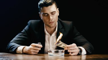 Businessman holding burning dollar and lighter near ashtray on table isolated on black 