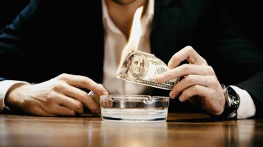 Cropped view of businessman holding burning money and lighter near ashtray isolated on black 