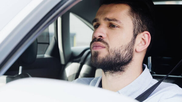 young and concentrated man looking ahead while driving car on blurred foreground