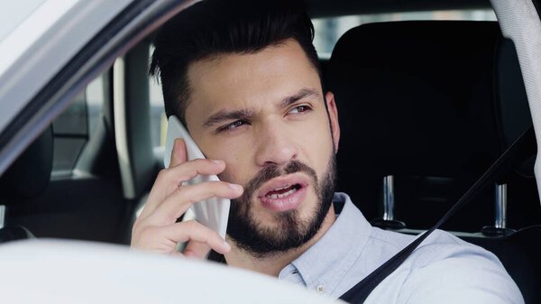 young bearded man driving car while looking away and talking on smartphone