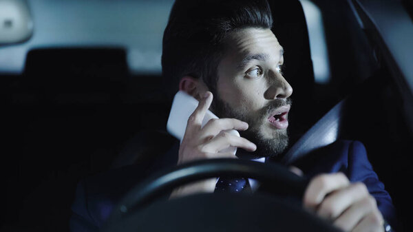 shocked businessman talking on smartphone and looking aside while driving car at night
