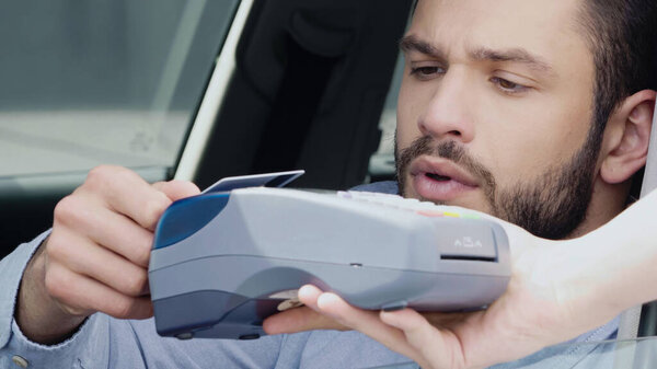 young bearded man holding credit card near payment terminal while sitting in car